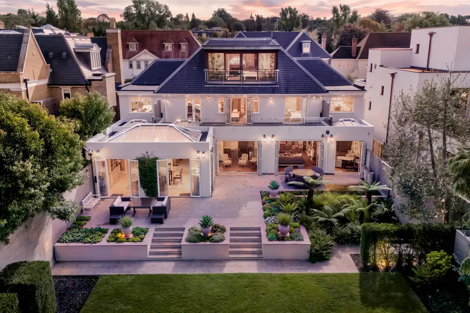 Rear elevation of a detached house at dusk, lit from inside, with terraced garden and balconies